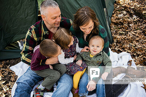 Smiling family looks at baby sitting on blanket in Fall leaves campin