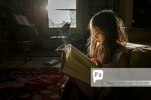 A small child sits in patch of bright sunlight reading a large book