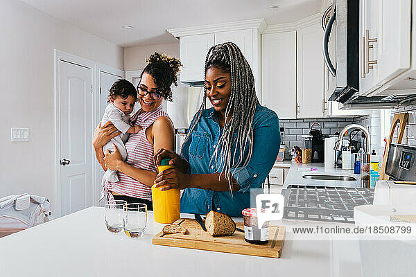 Happy mother carrying baby boy while girlfriend preparing breakfast in kitchen