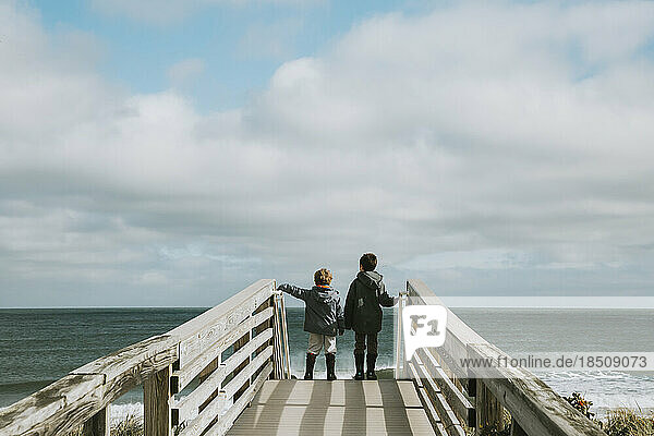 Two boys on boardwalk leading to beach against ocean and cloudy sky