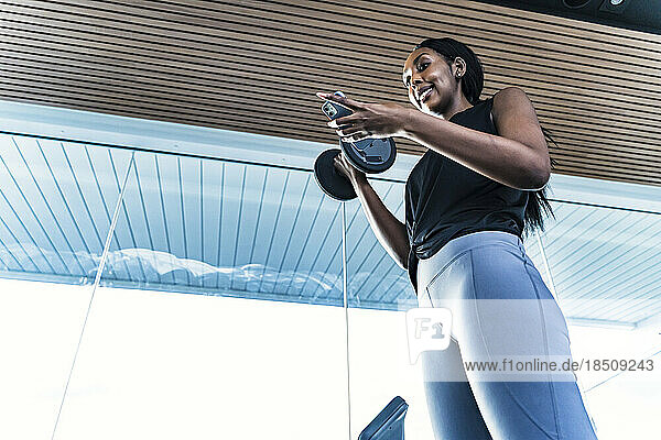 Young black girl in the gym doing exercises with a barbell and phone