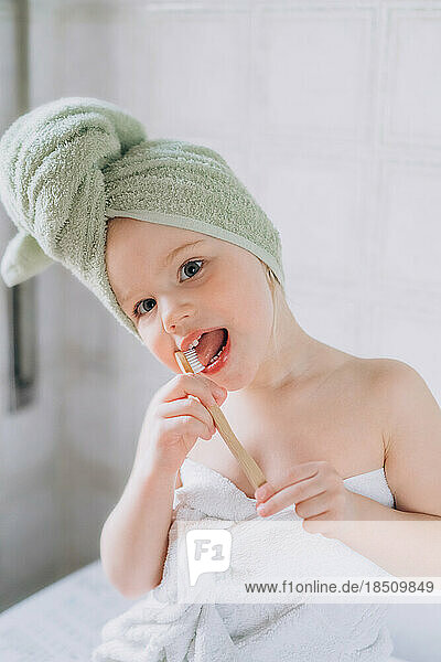 Toddler baby with a towel on her head brushes teeth with brush