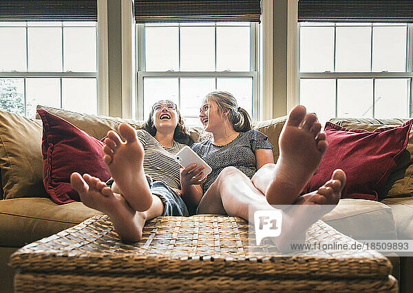 Two teenage girls with phone sitting on couch with feet up laughing.
