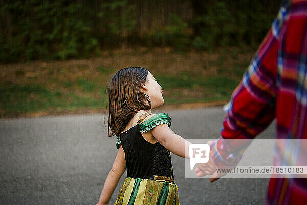 A small child in a princess costume holds her father's hands on a walk