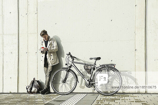 Young man text messaging on mobile phone and leaning against concrete wall  Munich  Bavaria  Germany