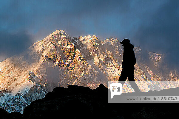 A man walks in silhouette on a ridge with Nuptse in the Himalayan background at sunset.