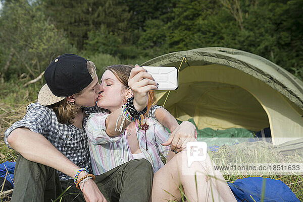 Young couple kissing and taking selfie with smart phone in front of camp tent  Bavaria  Germany