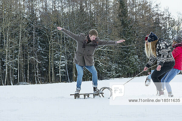 Three girls pulling young man standing on sled in snowy landscape  Bavaria  Germany