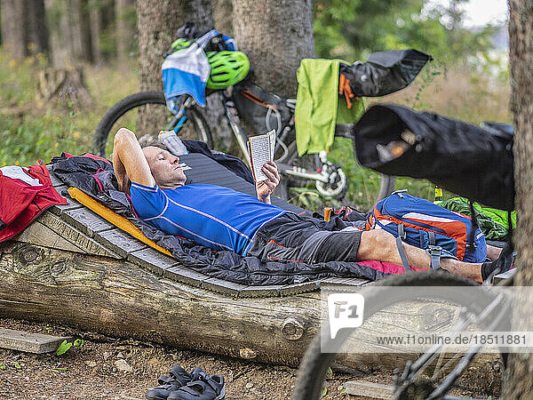Mountain biker resting on a log reading book amidst woods  Baden-Württemberg  Germany