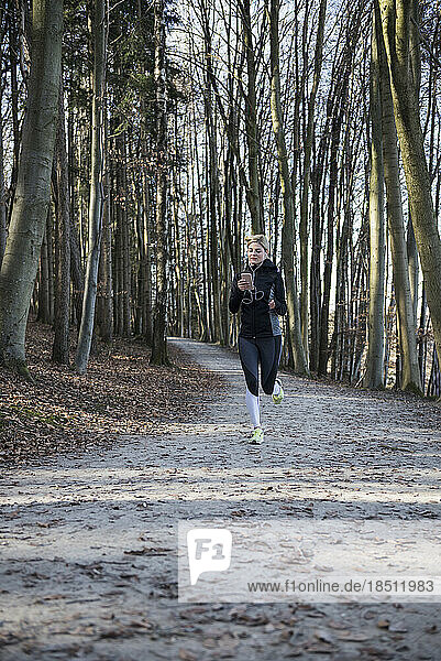 Fit woman with smart phone and headphones jogging in forest