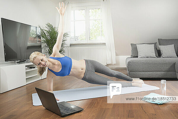 Young woman with laptop doing side plank pose on exercise mat in living room  Bavaria  Germany