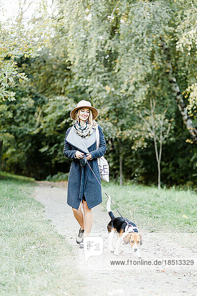 young woman in a coat and hat walks a beagle dog in the park