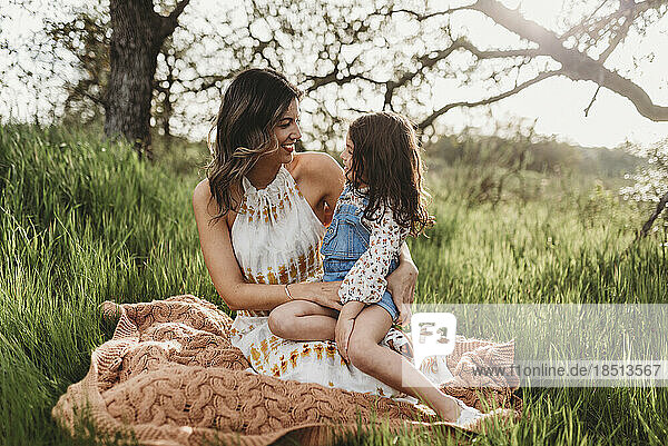 Mother and daughter sitting on blanket in field smiling at each other