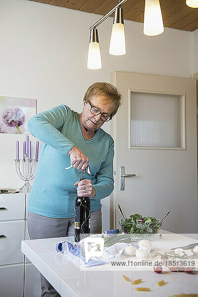Old woman opening a bottle of red wine in the kitchen