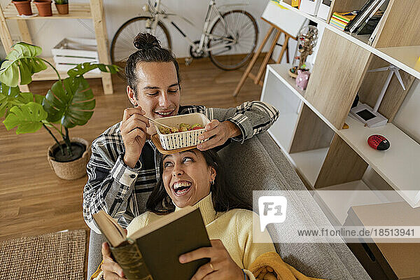 Smiling man eating snack with girlfriend reading book lying on sofa at home