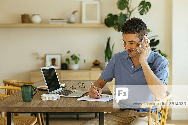 Smiling freelancer talking on mobile phone and taking notes on paper at table