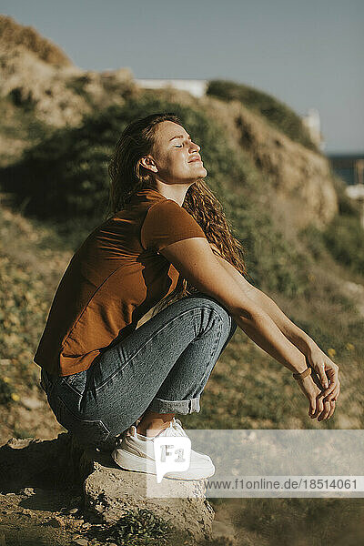 Woman with eyes closed crouching on rock at sunny day