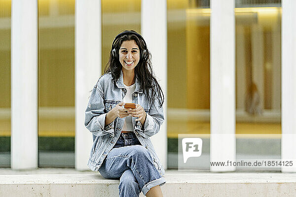 Glückliche Frau mit kabellosen Kopfhörern sitzt mit Smartphone vor dem Gebäude