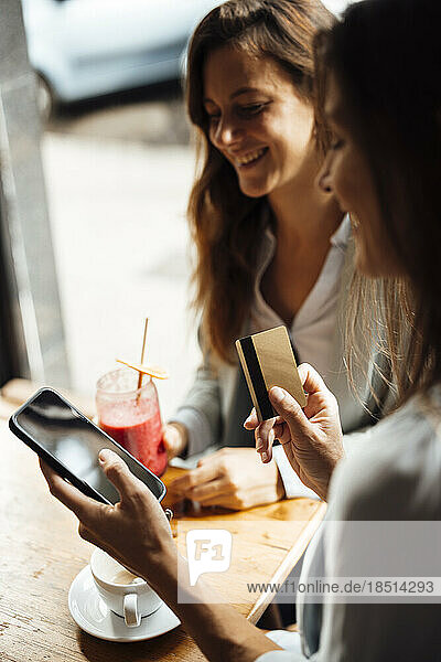 Woman holding credit card using smart phone by friend in cafe