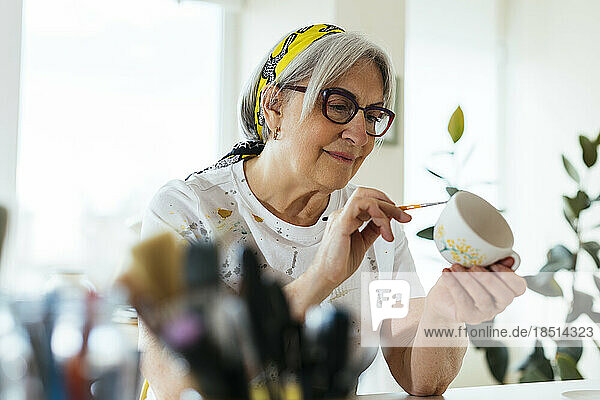Smiling woman wearing eyeglasses painting ceramic cup in workshop