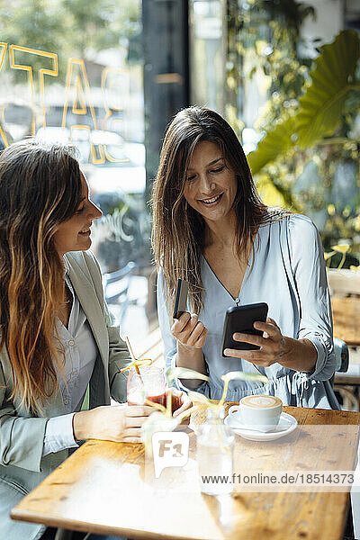 Woman with friend holding credit card using smart phone in cafe