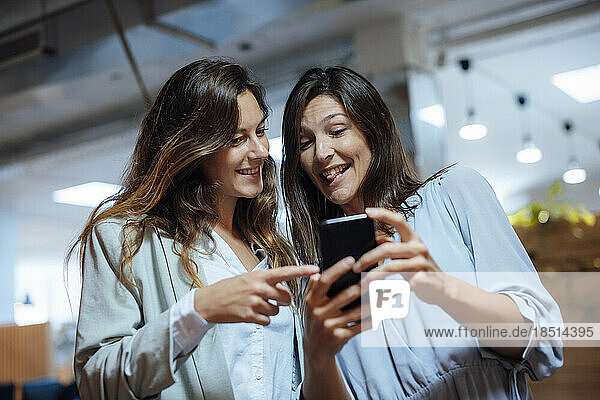 Happy businesswoman sharing mobile phone with colleague in office