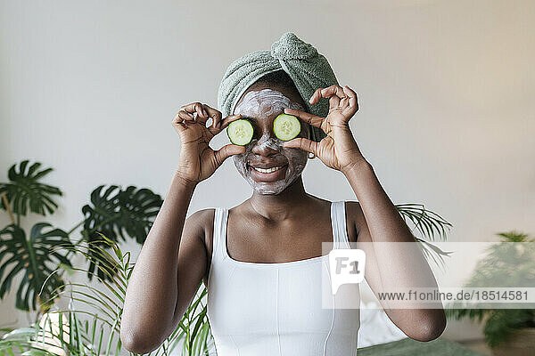 Smiling woman holding cucumber over eyes at home