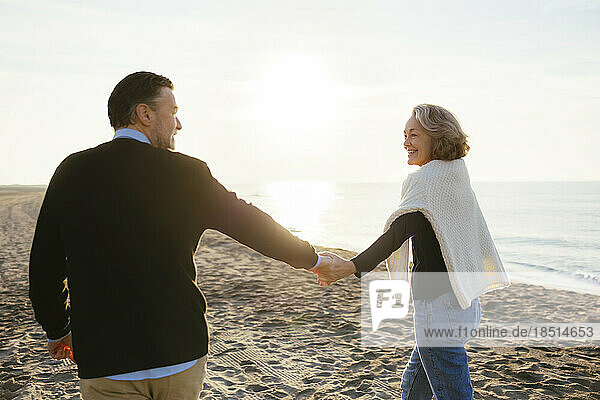 Smiling mature woman holding hands with man and walking at beach