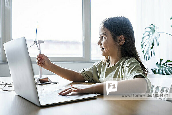 Girl with laptop touching wind turbine model on table at home