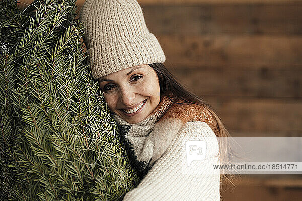 Glückliche Frau mit Strickmütze umarmt den Weihnachtsbaum