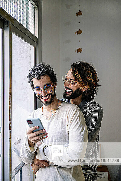 Happy gay man embracing boyfriend using smart phone near window at home