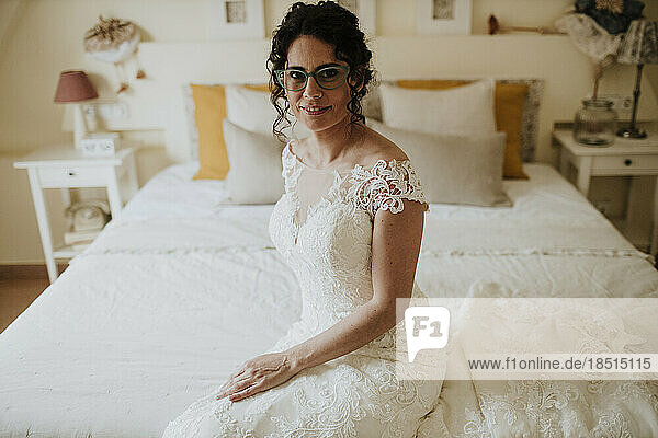 Lächelnde Braut im Hochzeitskleid  die auf dem Bett sitzt