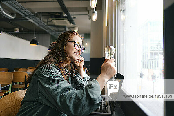 Smiling young freelancer looking at light bulb sitting by window in cafe