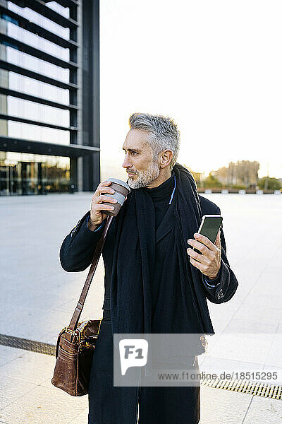 Mature businessman standing with coffee cup and smart phone at footpath
