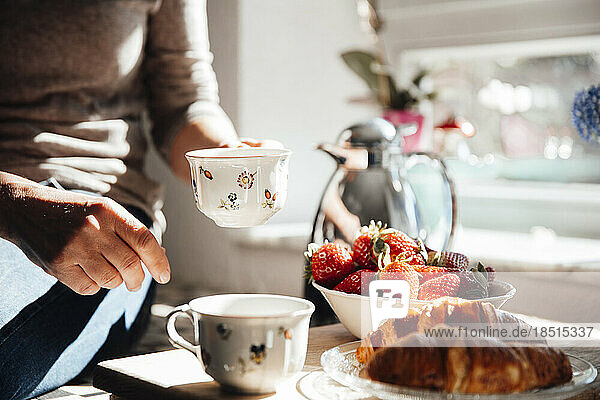 Woman holding coffee cup with croissant and fresh strawberries in kitchen