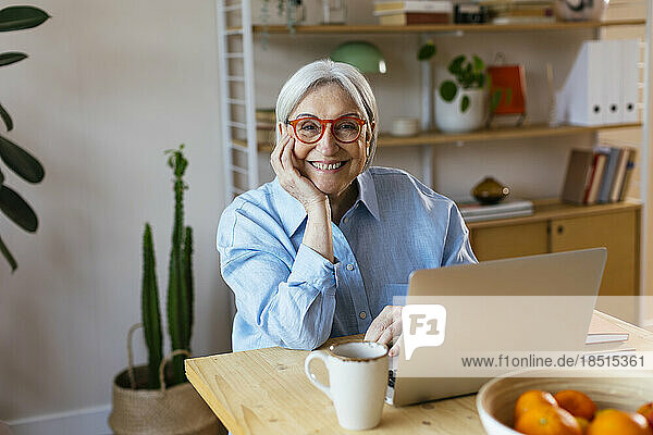 Happy senior woman sitting with hand on chin at home