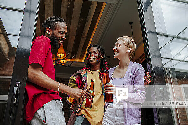 Happy multiracial friends enjoying beer at doorway of apartment