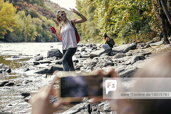 Daughter photographing mother showing peace sign standing near river