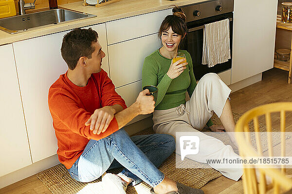 Happy woman having juice with man holding coffee cup sitting by cabinet in kitchen