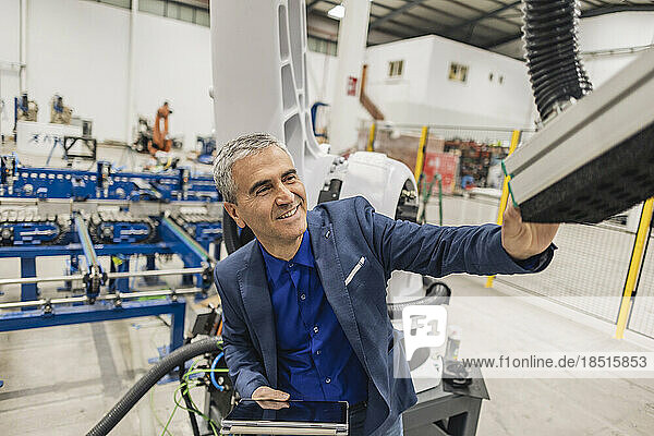 Smiling engineer with tablet PC touching robotic arm in industry