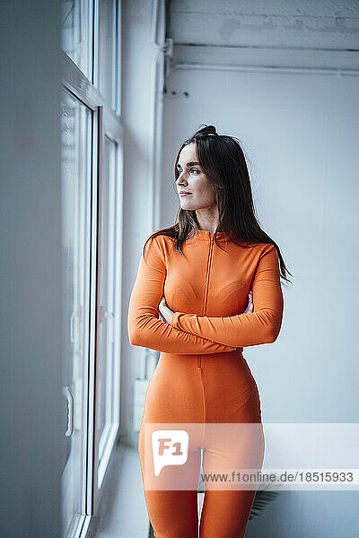 Young businesswoman in orange jumpsuit standing with arms crossed by window in office