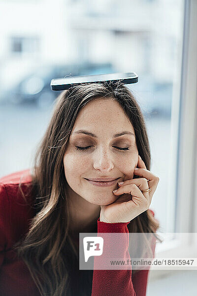 Woman with eyes closed balancing smart phone on head in front of window