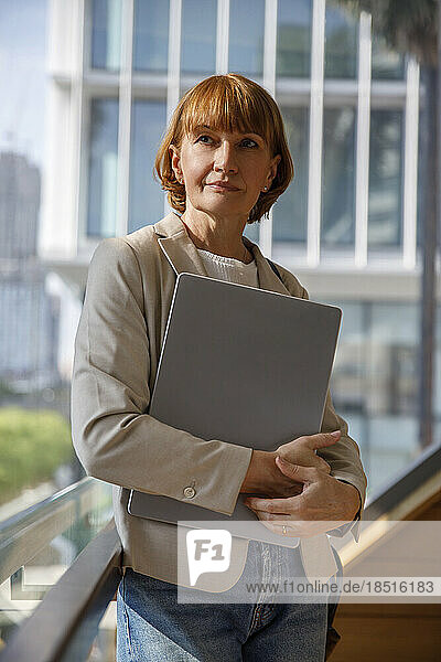 Thoughtful mature businesswoman holding laptop in front of window