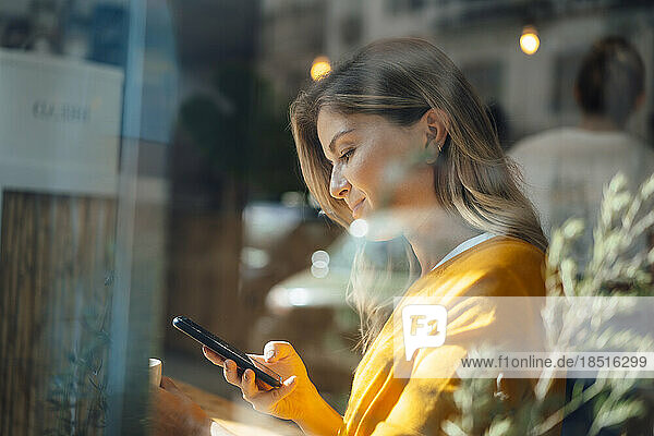 Smiling woman using smart phone in cafe seen through glass
