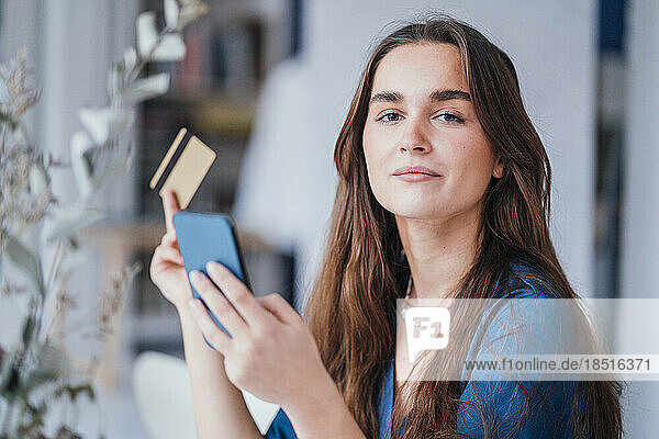 Confident young businesswoman with mobile phone and credit card in office