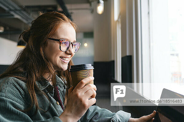Happy young freelancer with disposable coffee cup sitting in cafe