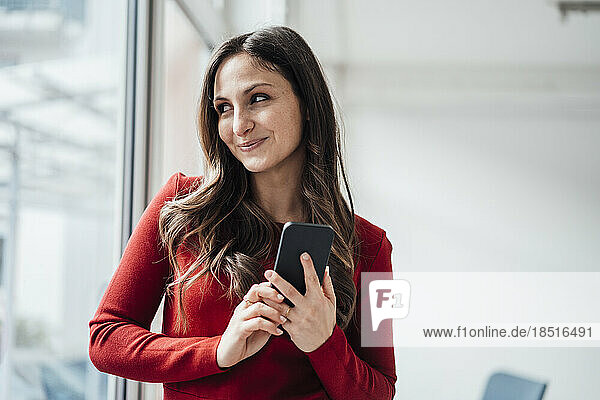 Smiling businesswoman with smart phone looking through window