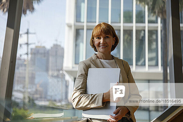 Smiling mature businesswoman with laptop in front of window