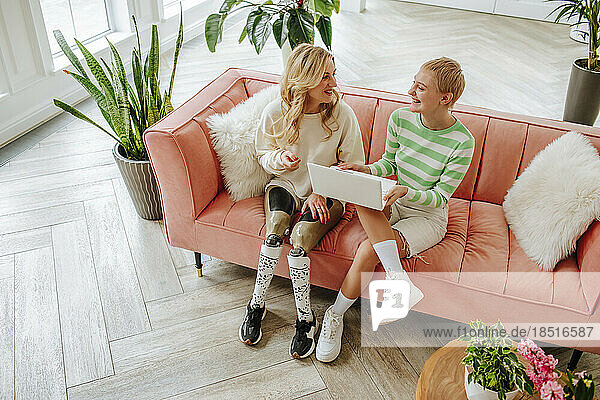 Woman with disability sitting by friend holding laptop in living room at home