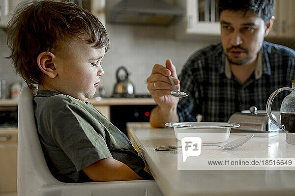 Father feeding upset son sitting at dining table in home